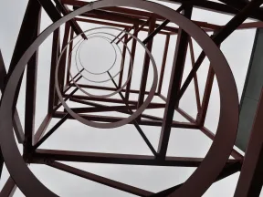 Looking upward inside a red metal tower structure with circular and diagonal beams against a cloudy sky.