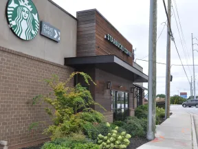 Starbucks coffee shop exterior with drive-thru sign and landscaped greenery by busy street corner.