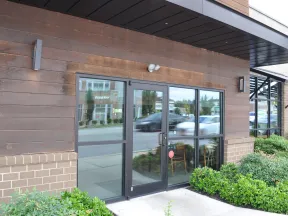Glass double doors with black frames at storefront reflecting street and cars, surrounded by wood and brick exterior with greenery.
