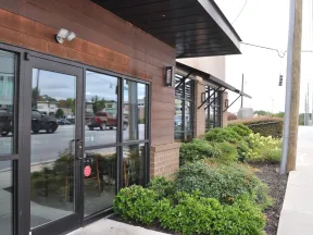 Commercial building entrance with glass doors and landscaped greenery along the sidewalk on a busy street.