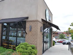 Exterior view of a Starbucks coffee shop with brick walls, large windows, awnings, and surrounding greenery on a cloudy day.