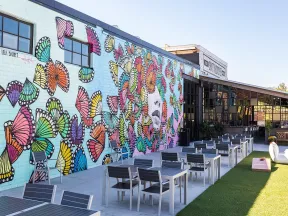 Outdoor patio with modern tables and chairs next to a colorful butterfly mural on a brick wall under clear sky