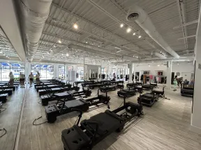 Spacious Pilates studio with multiple reformer machines, light wood floors, and people preparing for class.