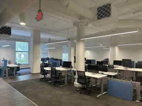 Modern office space with multiple desks, black chairs, and computer monitors under bright lighting and large windows.