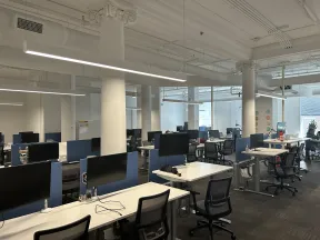 Modern open office with rows of desks, computer monitors, office chairs, and white columns under bright lighting.