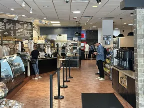 Spacious cafe interior with customers ordering, menu boards, snack shelves, and coffee station with tiled floor.