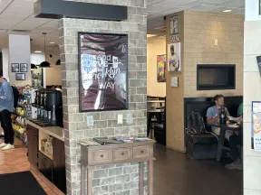 Interior of a cafe with brick walls, framed Brooklyn poster, coffee station, and a man dining at a booth.