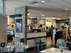 Interior of a casual café with customers ordering at counter, tables and chairs, and a visible menu board.