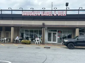 Brooklyn Bagel & Deli storefront with outdoor seating and customers under cloudy sky in a shopping plaza.