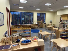 Montessori classroom with wooden tables, chairs, educational materials, and large windows letting in natural light