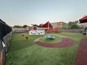 Empty modern playground with artificial turf, red and gray play elements, and a shaded canopy near buildings.