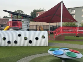 Modern playground with a large red shade canopy, corrugated tunnel, colorful spinning play equipment, and a slide.
