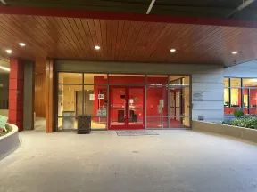 Entrance to a modern building with red doors, glass panels, wooden ceiling, and concrete floor at dusk