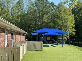 Outdoor playground with blue shade sails, artificial grass, play structures, and fenced area next to brick building.