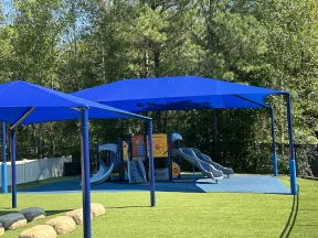 Outdoor playground with blue slides, climbing structures, and blue shade canopies surrounded by greenery.