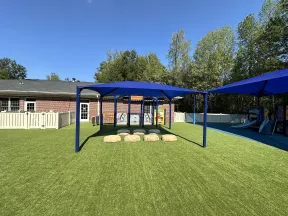 Playground area with artificial grass, blue shaded canopies, stepping stones, and a brick building in background