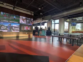 Modern sports bar with large screens showing games, industrial ceiling, wooden benches, and people near the bar counter.