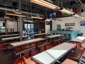 Modern industrial-style cafeteria with long tables, bench seating, hanging lights, and a bar area in the background