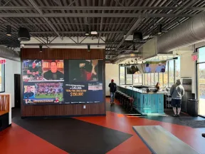 Modern sports bar interior with large TV wall, blue bar counter, industrial ceiling, and customers inside.