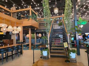 Modern indoor restaurant with wooden tables, hanging lights, plants, staircase, and arcade games in the background