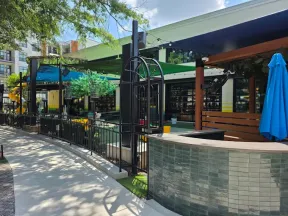 Outdoor patio area with shaded seating, black metal fencing, tiled counter, and blue umbrella on a sunny day.