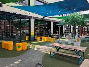 Outdoor seating area with picnic tables, yellow recycling bins, shade sails, and string lights in a commercial space.