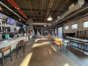 Spacious brewery tasting room with bar stools, wooden tables, industrial ceiling, and wall art of cows and text.