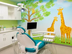 Colorful children's dental office with a dental chair and wall mural of giraffes and a tree with an owl.