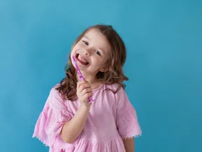 Smiling young girl in pink shirt holding a purple toothbrush against a blue background