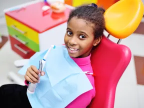 Smiling young girl sitting in a colorful dental chair holding an electric toothbrush at the dentist office.
