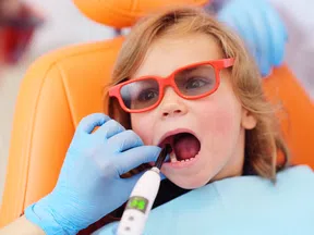 Child wearing red glasses receiving dental treatment with dental curing light in orange dental chair.