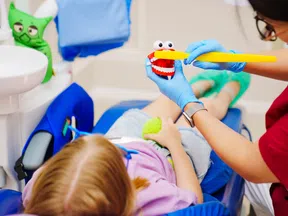 Dentist demonstrates proper brushing on a toy mouth to a child patient lying in the dental chair.