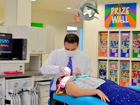 Dentist treating a young girl wearing pink headphones in a bright pediatric dental clinic with a colorful prize wall.