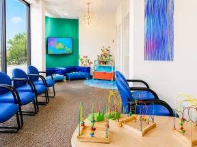 Bright pediatric waiting room with blue chairs, children's toys, colorful wall art, and large windows with natural light.