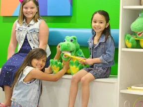 Three smiling girls in colorful clothes play with a large green crocodile toy against a bright green wall.