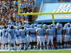 University football team in light blue and white uniforms gathers on the field near the goalpost with crowd in stands.