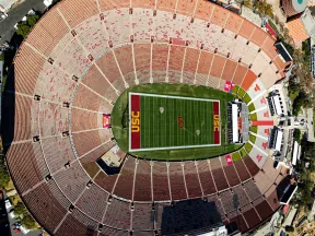 Aerial view of the USC football stadium showing empty seats and the green field with USC logos.