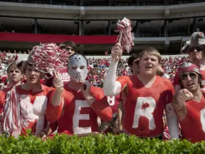 College football fans with painted red and white bodies cheering in a crowded stadium during a game.