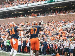 Two football players in orange and blue uniforms celebrate a touchdown in front of a cheering crowd in a stadium.