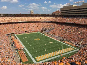 Aerial view of a packed football stadium with orange and white crowd and players on the field under a blue sky.
