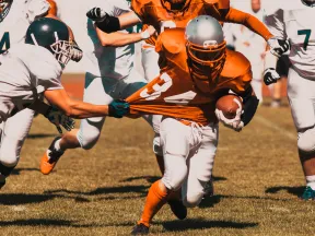 A football player in orange evades tackles while running with the ball during a game.