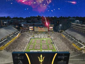 Fireworks explode above a full stadium with green field and large digital scoreboard at dusk.