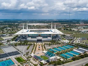 Aerial view of Hard Rock Stadium surrounded by parking lots and tennis courts under a cloudy sky.