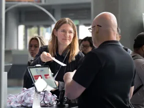 Smiling woman hands a ticket to a bald man at a service counter with people waiting behind them.