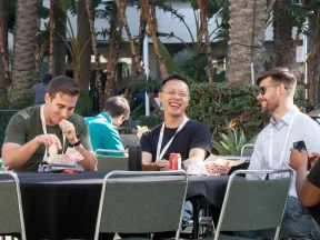 Four men sitting at a table outdoors, eating, drinking, and enjoying a sunny social gathering with palm trees in the background.