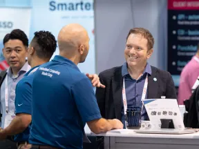 Business professionals engaged in conversation at a technology trade show booth with product displays and banners.