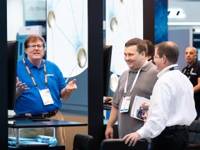 Three men wearing conference badges engaged in discussion at a technology trade show booth.
