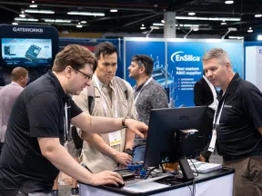 Three men engaged in a tech demonstration at a conference booth with computer and electronic components.