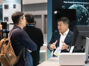 Business professionals engaged in discussion at a technology trade show booth with computer displays.