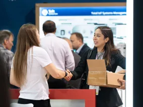 Two professional women shaking hands at a conference booth with a crowd and display screen in background
